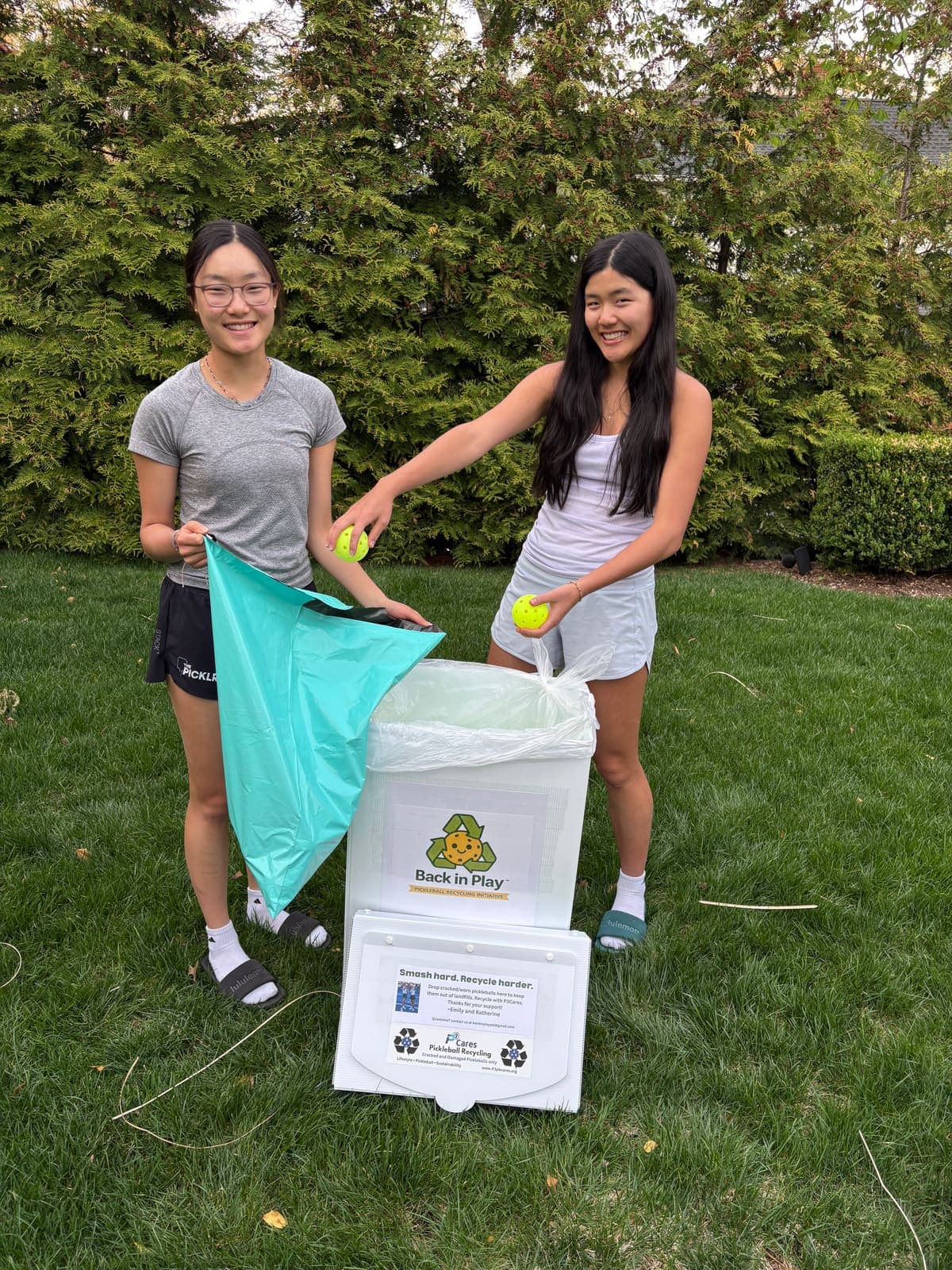 Twin sisters Emily and Katherine dropping cracked pickleballs into a Back in Play collection bin on a grass lawn