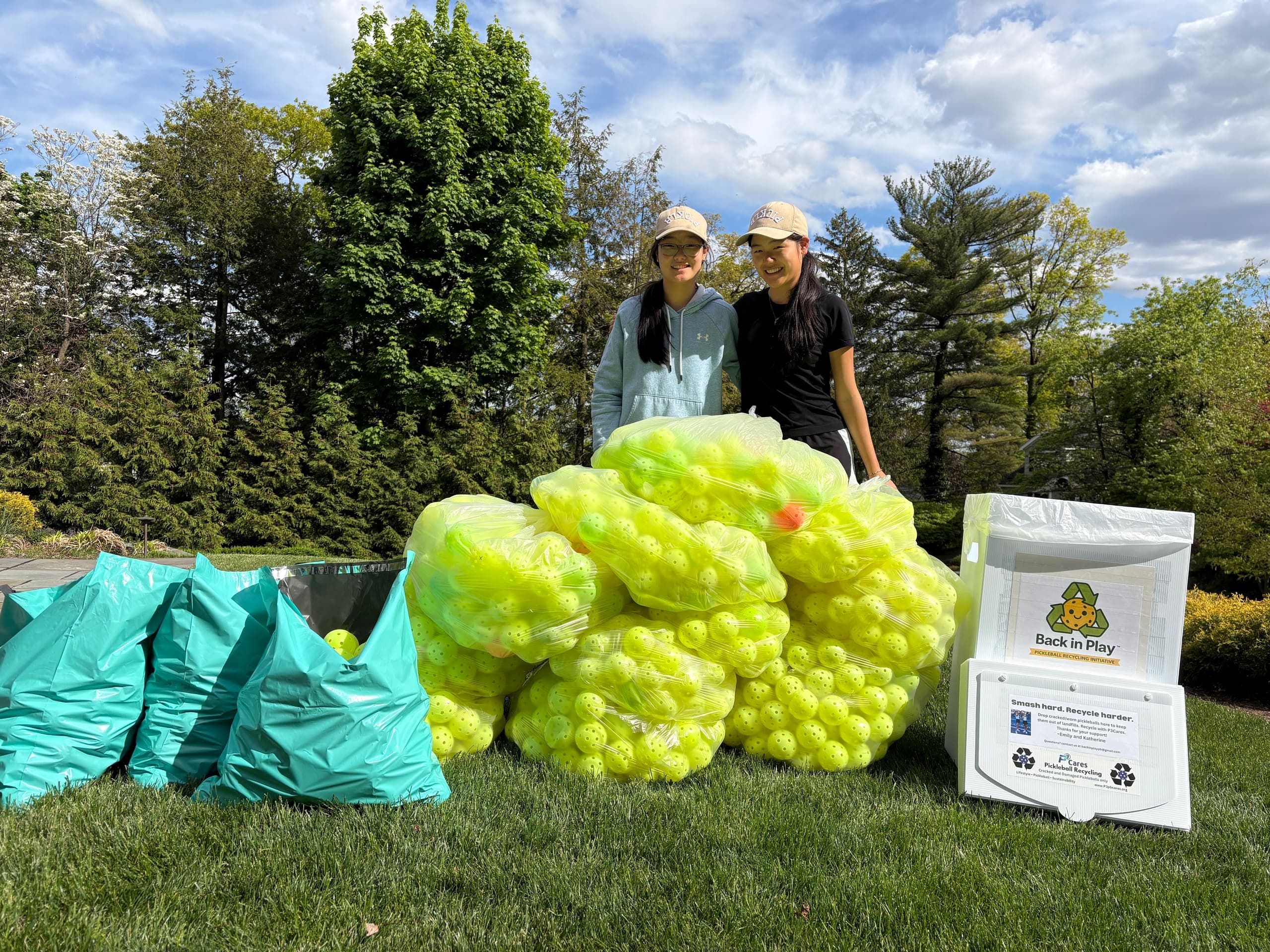 Twin founders Emily and Katherine standing behind a large pile of bagged, collected pickleballs next to a branded Back in Play collection bin